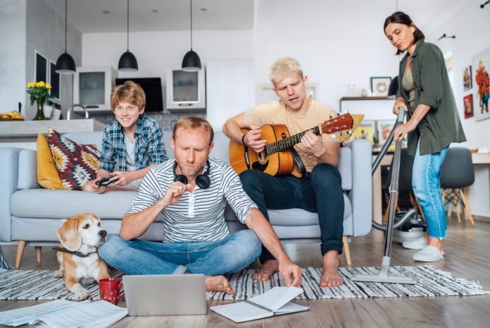 Quarantine time: Family at living room together. Father distance working using laptop, mother cleaning the room, older son playing acoustic guitar and singing a songs, little son playing video console