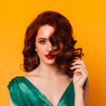 Front view of sensual woman with ginger wavy hair. Studio shot of elegant girl in green dress isolated on yellow background